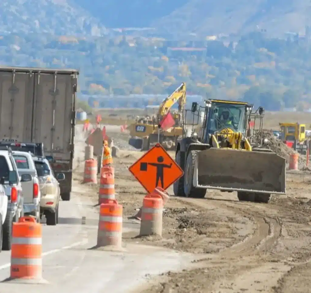 Highway roadwork zone with heavy equipment, traffic barrels, and vehicles moving through a controlled construction area