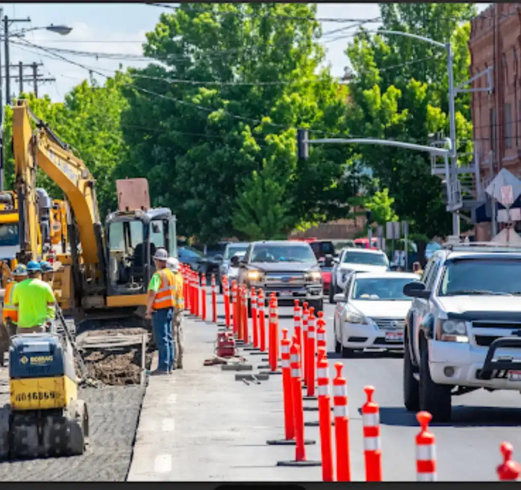 Urban road construction crew managing traffic with delineators and heavy equipment beside active vehicle lanes